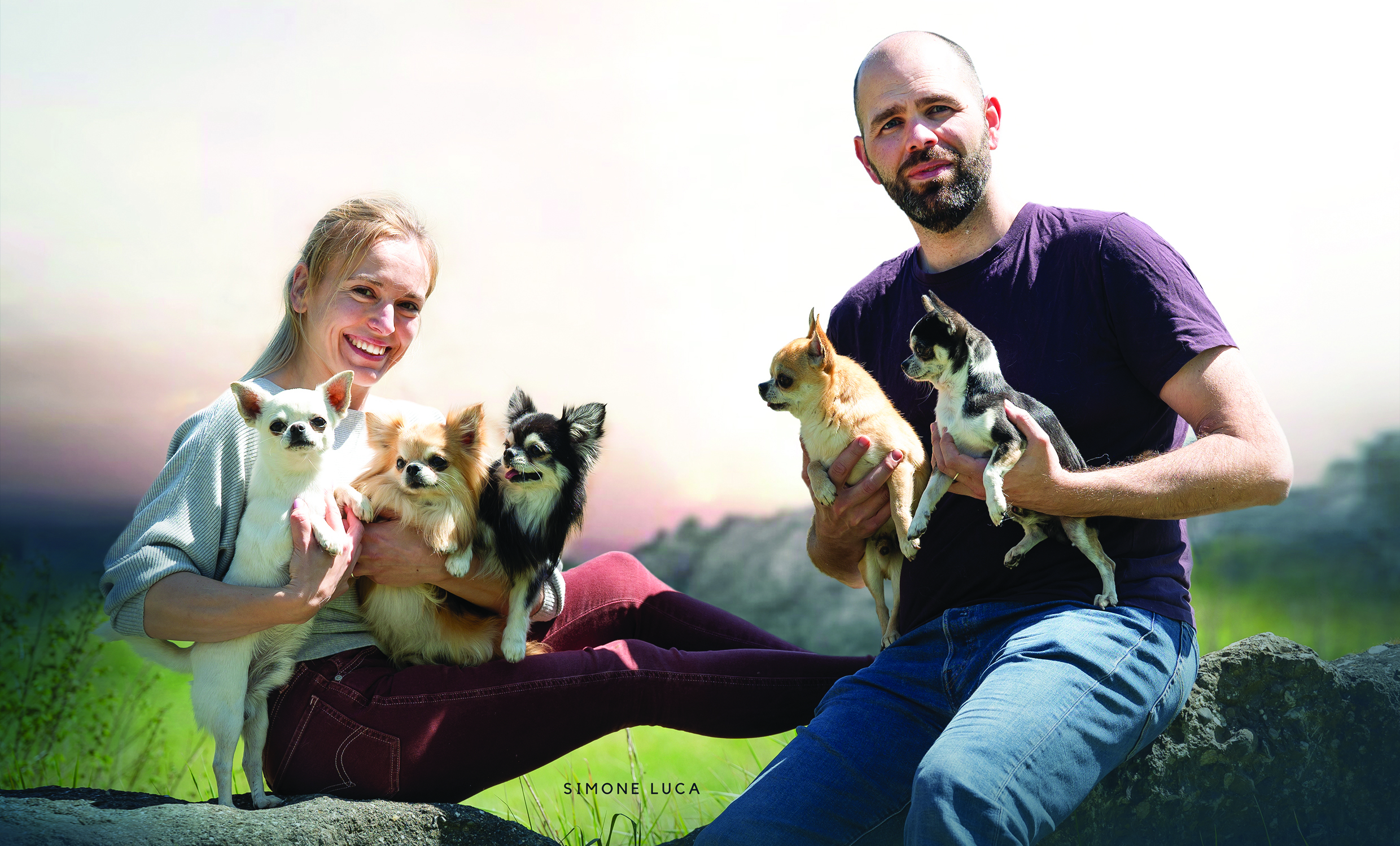 A couple outdoors posing with multiple small dogs on a rocky ledge, clear sunny sky behind them.